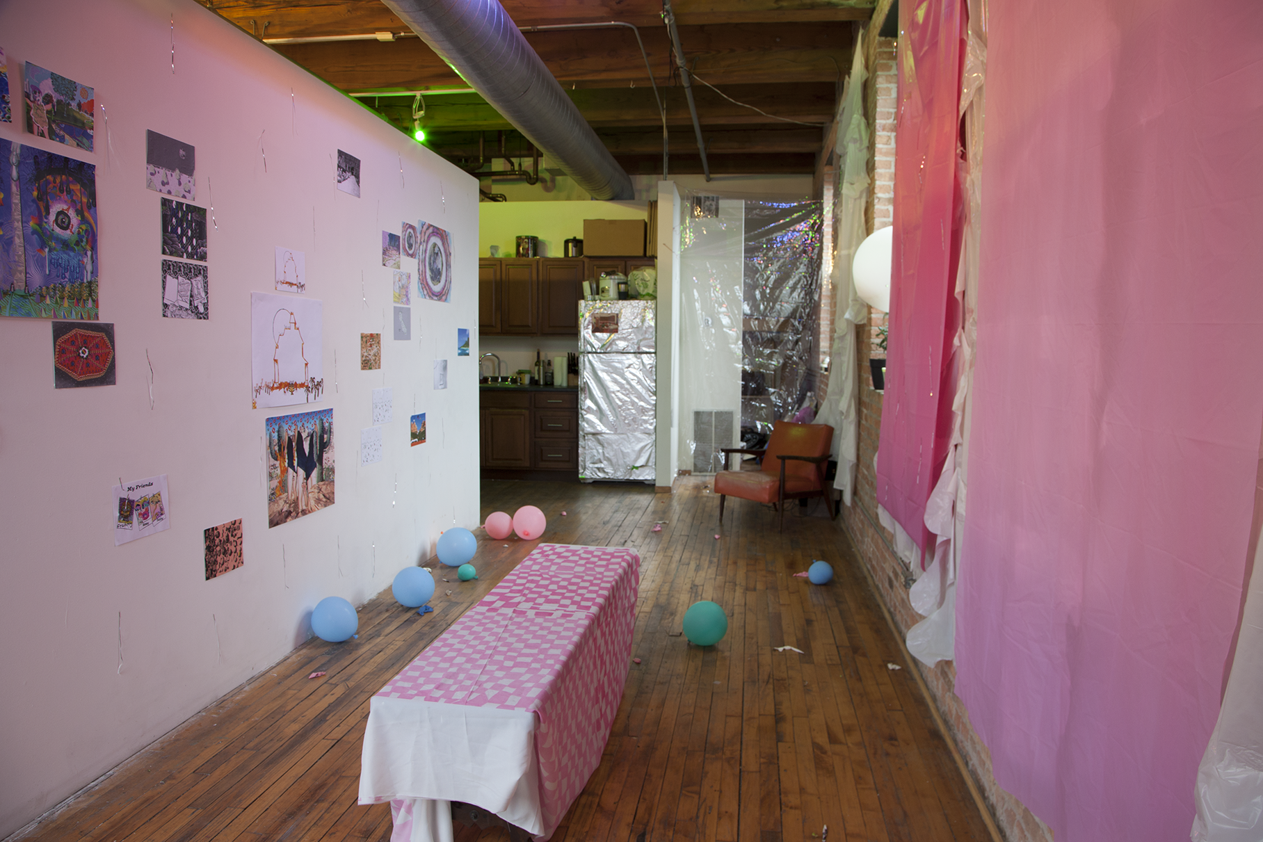 Installation view of many small scale prints hung on white walls on the left and many pink curtains and baloons on the right